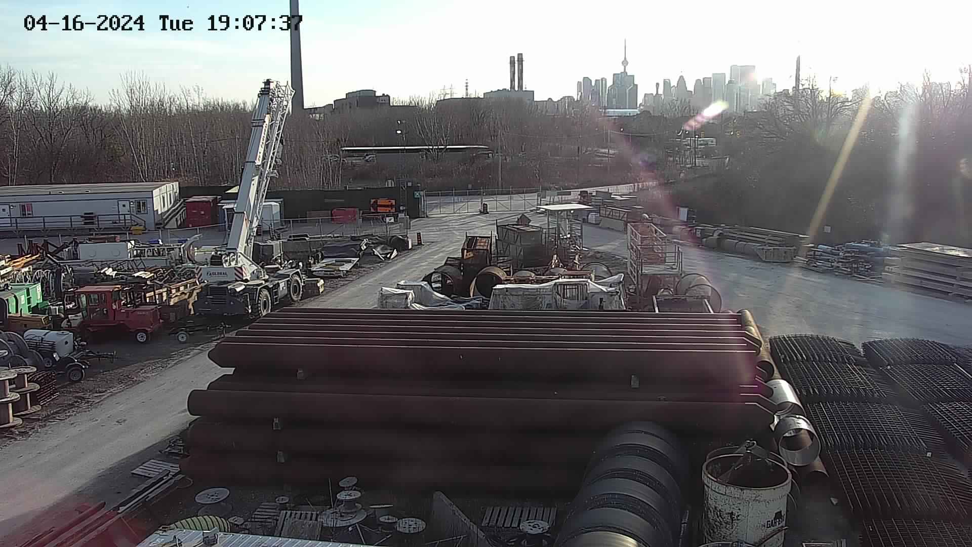 LSS camera view over an industrial laydown yard at dusk, with a crawler crane and stacked pipe, timestamped by the camera